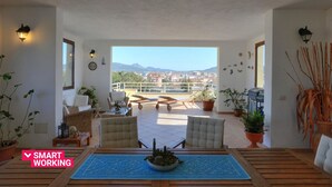 Un vaste balcon offrant une vue sur le paysage urbain et les montagnes, aménagé avec des chaises et une table, et agrémenté de plantes et d’œuvres d’art murales.