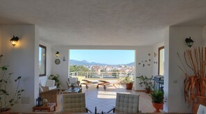 Un vaste balcon offrant une vue sur le paysage urbain et les montagnes, aménagé avec des chaises et une table, et agrémenté de plantes et d’œuvres d’art murales.