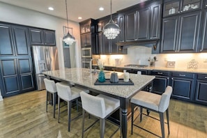 A modern kitchen with dark cabinetry, a granite island, bar stools, stainless steel appliances, and pendant lighting.