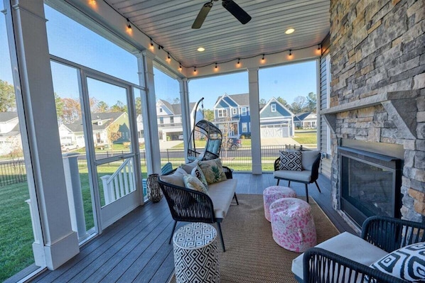 A covered porch with a stone wall, a fireplace, outdoor seating, and a view of houses in the background.