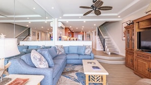 A spacious living room with a large sectional sofa, a coffee table, and a television on a wooden cabinet. A staircase leads to an upper level. - New Unit To rental Program (Lahaina)