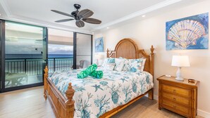 A bedroom with a bed, a ceiling fan, a nightstand, a painting, and a view of the ocean. - New Unit To rental Program (Lahaina)