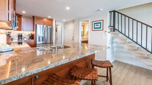 A kitchen with granite countertops, stainless steel appliances, and wooden bar stools. - New Unit To rental Program (Lahaina)
