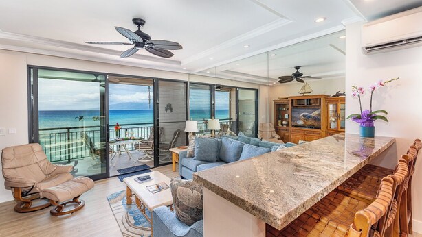 A spacious living area with a large sliding glass door leading to a balcony with a view of the sea. There is a comfortable seating area with a sofa and chairs, a kitchen counter, and a ceiling fan. - New Unit To rental Program (Lahaina)