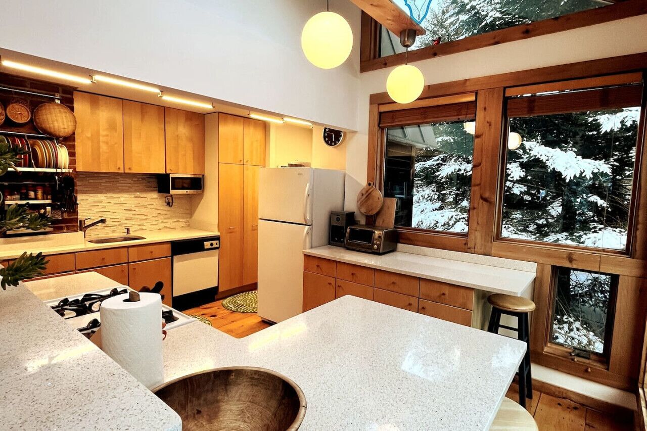 A modern kitchen with wooden cabinets, a large island, and a view of snow-covered trees through large windows.