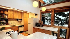 A modern kitchen with wooden cabinets, a large island, and a view of snow-covered trees through large windows. - Chalet- Stream, A/C, Hot Tub, min to Lake and Ski (Whitingham)
