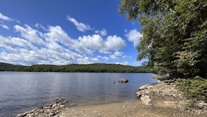 A rocky shoreline with a calm lake and a forested hill in the background. - Chalet- Stream, A/C, Hot Tub, min to Lake and Ski (Whitingham)