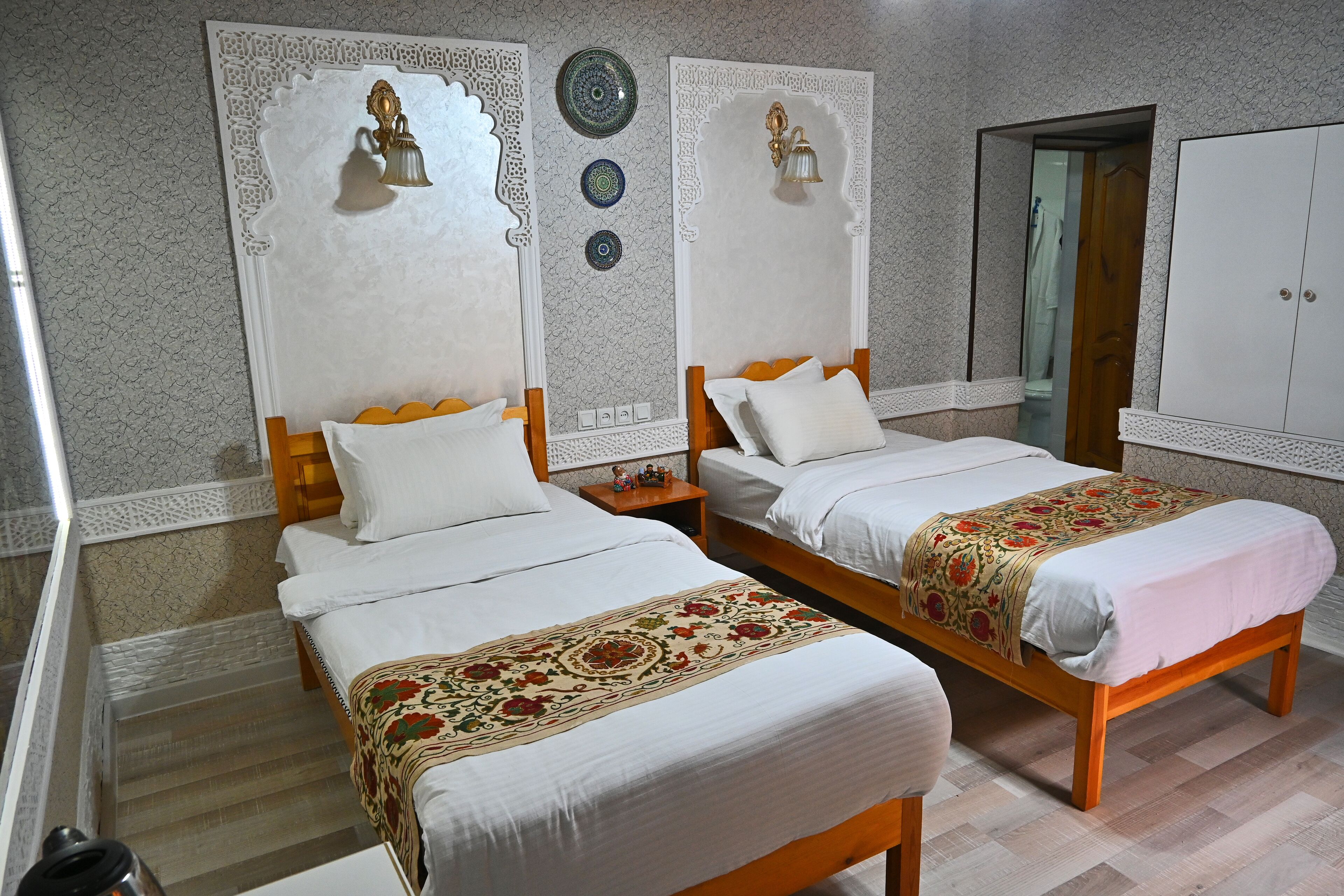 A hotel room with two beds, each with white bedding and decorative headboards. There are wall-mounted light fixtures and a wardrobe in the room.