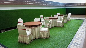 An outdoor seating area with round tables and chairs, surrounded by artificial grass, under a large overhang. - Hotel Saptarshi (Bankura)