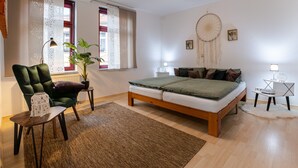 Une chambre à coucher avec un lit, une chaise, une table, une plante, un attrape-rêves et une fenêtre avec des rideaux.