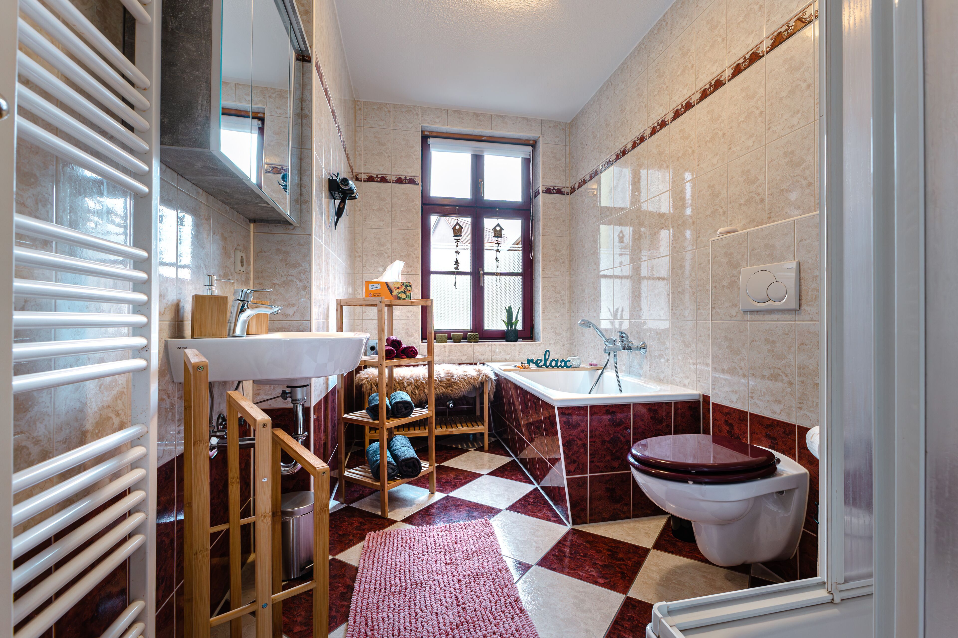 A bathroom with a checkered floor, a wooden shelf with towels, a sink, a toilet, and a window with decorative elements.