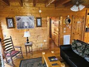 A cozy cabin interior with wooden walls, a rocking chair, a black leather sofa, a small table with a lamp, and a bear-themed wall hanging.