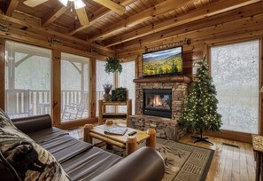 A cozy cabin interior with a stone fireplace, a television mounted above it, and a Christmas tree.
