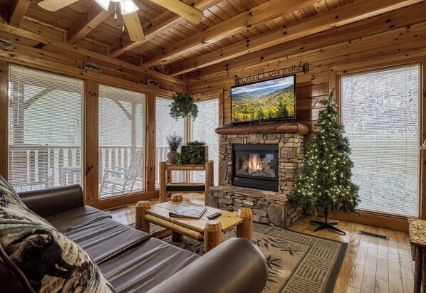A cozy cabin interior with a stone fireplace, a television mounted above it, and a Christmas tree.
