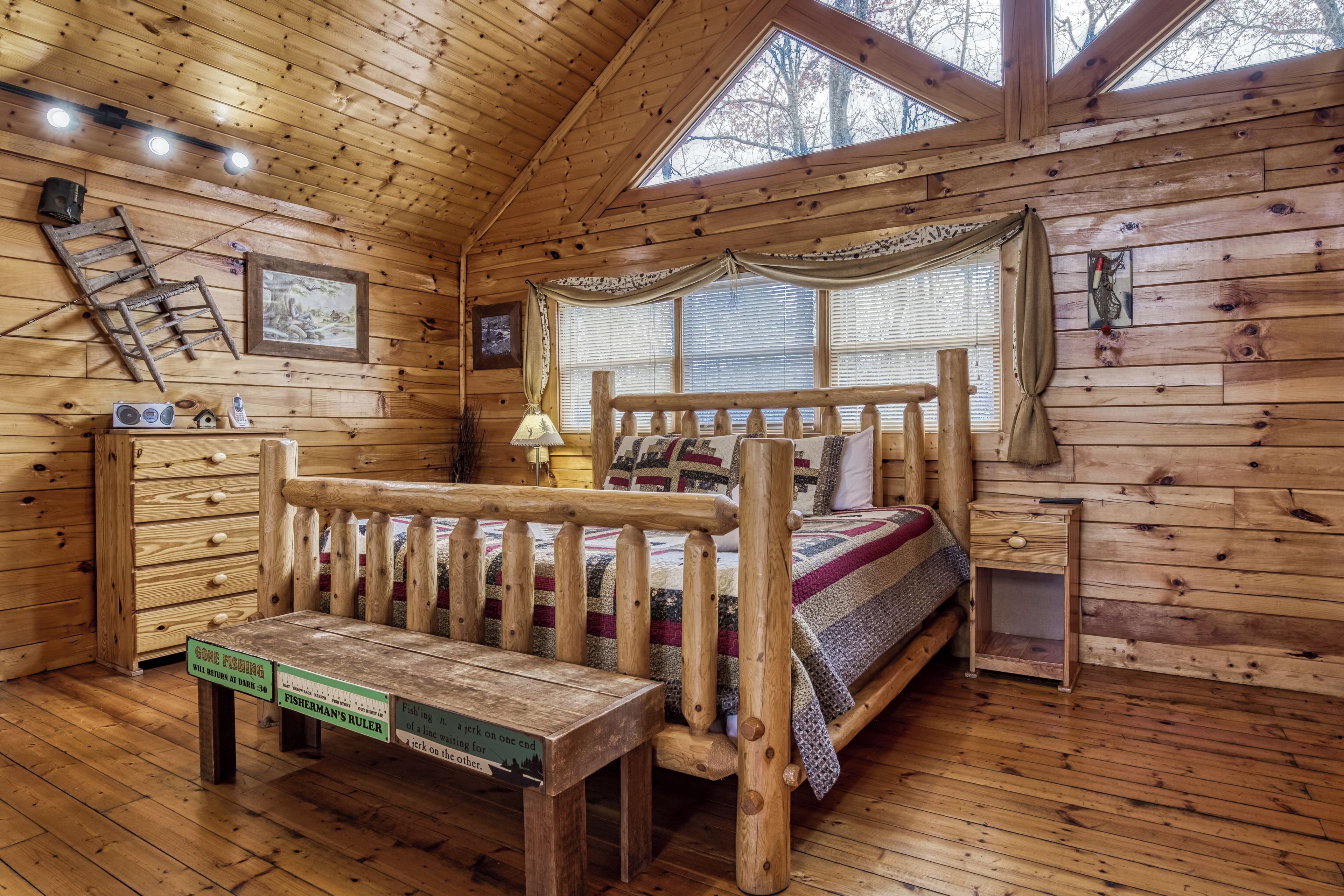 A wooden cabin interior with a bed, a bench, a dresser, and a window with blinds.