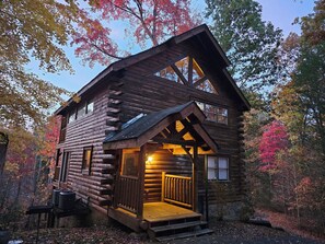 A log cabin with a porch and a small deck, surrounded by autumn foliage.