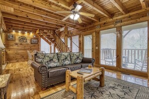 A wooden cabin interior with a leather sofa, a coffee table, and a staircase leading to an upper level.