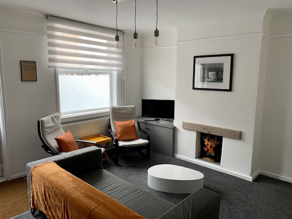 Ein modernes Wohnzimmer mit Sofa, Sessel und Couchtisch. Ein Kamin, ein gerahmtes Bild an der Wand und ein Fenster mit Jalousien sind ebenfalls vorhanden.