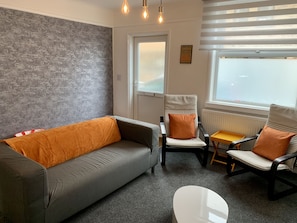 Ein modernes Wohnzimmer mit einem grauen Sofa, zwei Sesseln mit orangefarbenen Kissen, einem kleinen weißen Tisch und einem Fenster mit Jalousien.