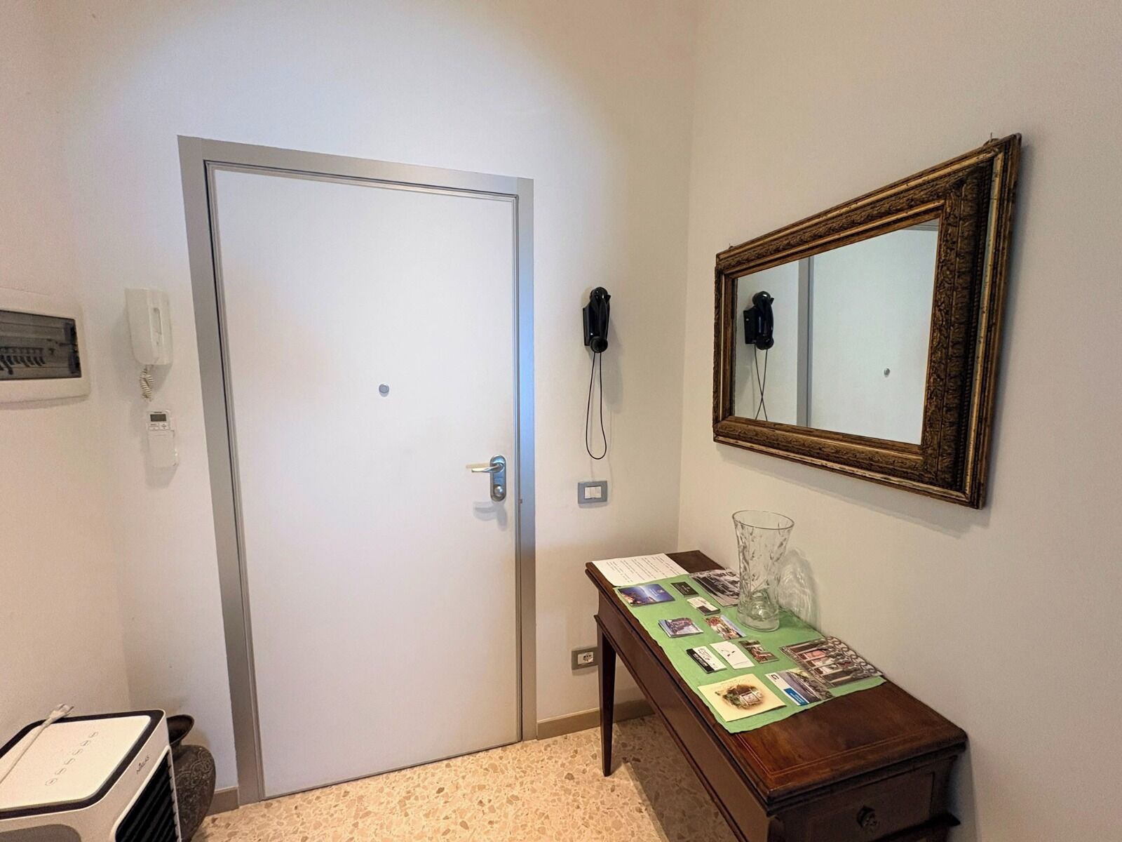 Une chambre d’hôtel avec une porte fermée, un téléphone, un miroir, une table avec un verre et un petit placard.