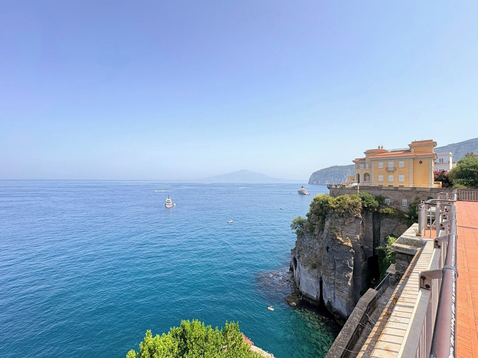 A coastal view with a building overlooking the sea, a cliff, and a clear sky.