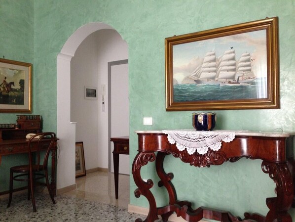 A room with a green wall, a framed painting of a sailing ship, a wooden table with a lace tablecloth, and a desk with a chair.