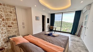 Une chambre avec un grand lit, un mur en pierre, une armoire, un bureau avec une chaise et un balcon avec vue.