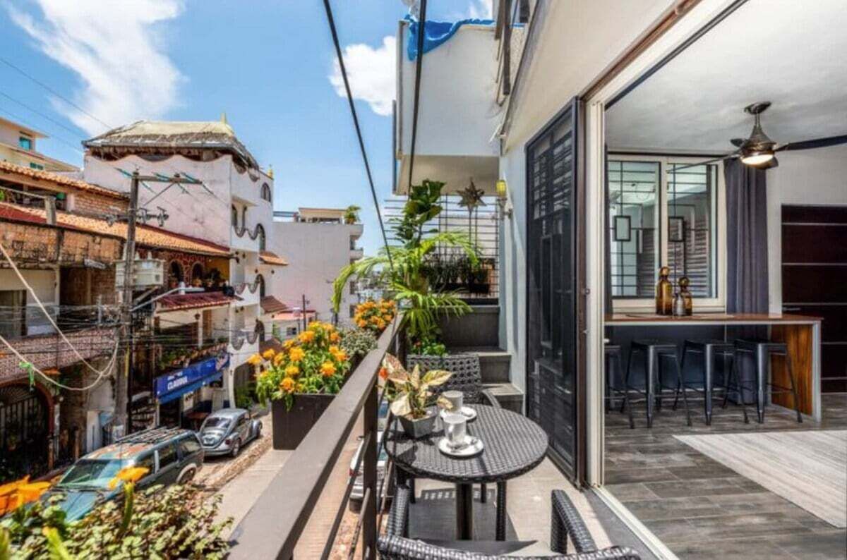 Een smal balkon in de stad met een tafeltje en stoelen, plantenbakken en uitzicht op omliggende gebouwen.