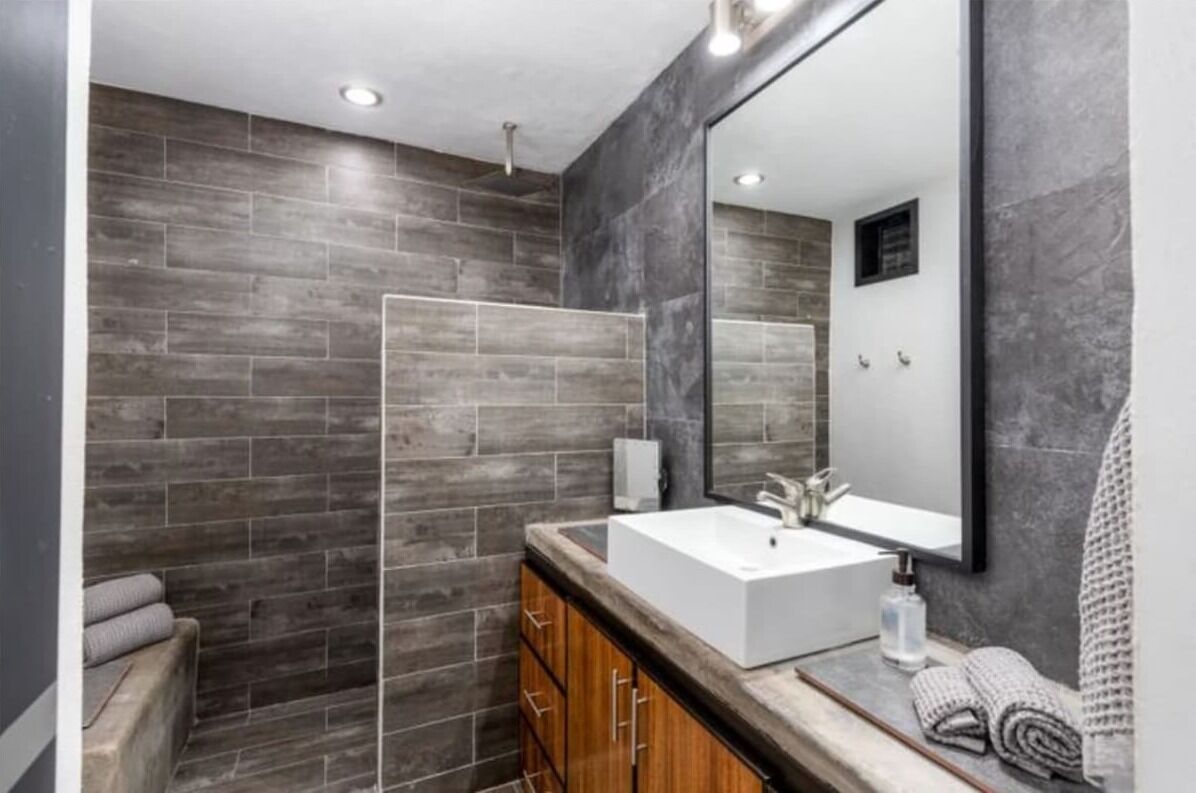Un baño moderno con un espejo grande, un lavabo blanco, armarios de madera y una zona de ducha con azulejos oscuros.