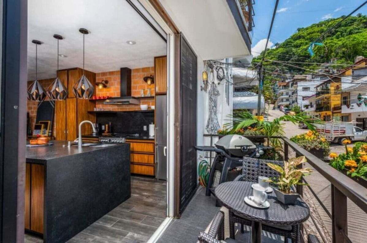 Una cocina moderna con encimera negra y armarios de madera, un balcón con mesa y sillas, y vistas a una zona residencial con casas y vegetación.