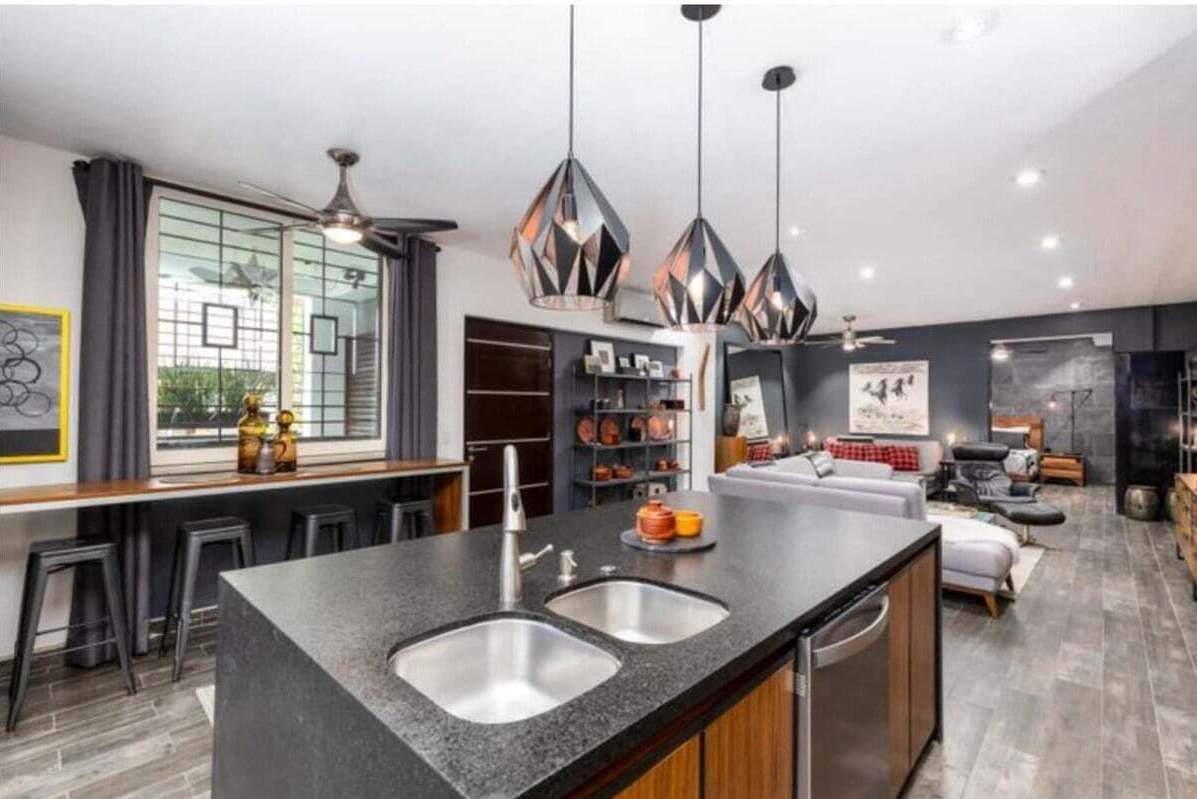 A modern kitchen with a central island, bar stools, pendant lighting, and a view of the living area.