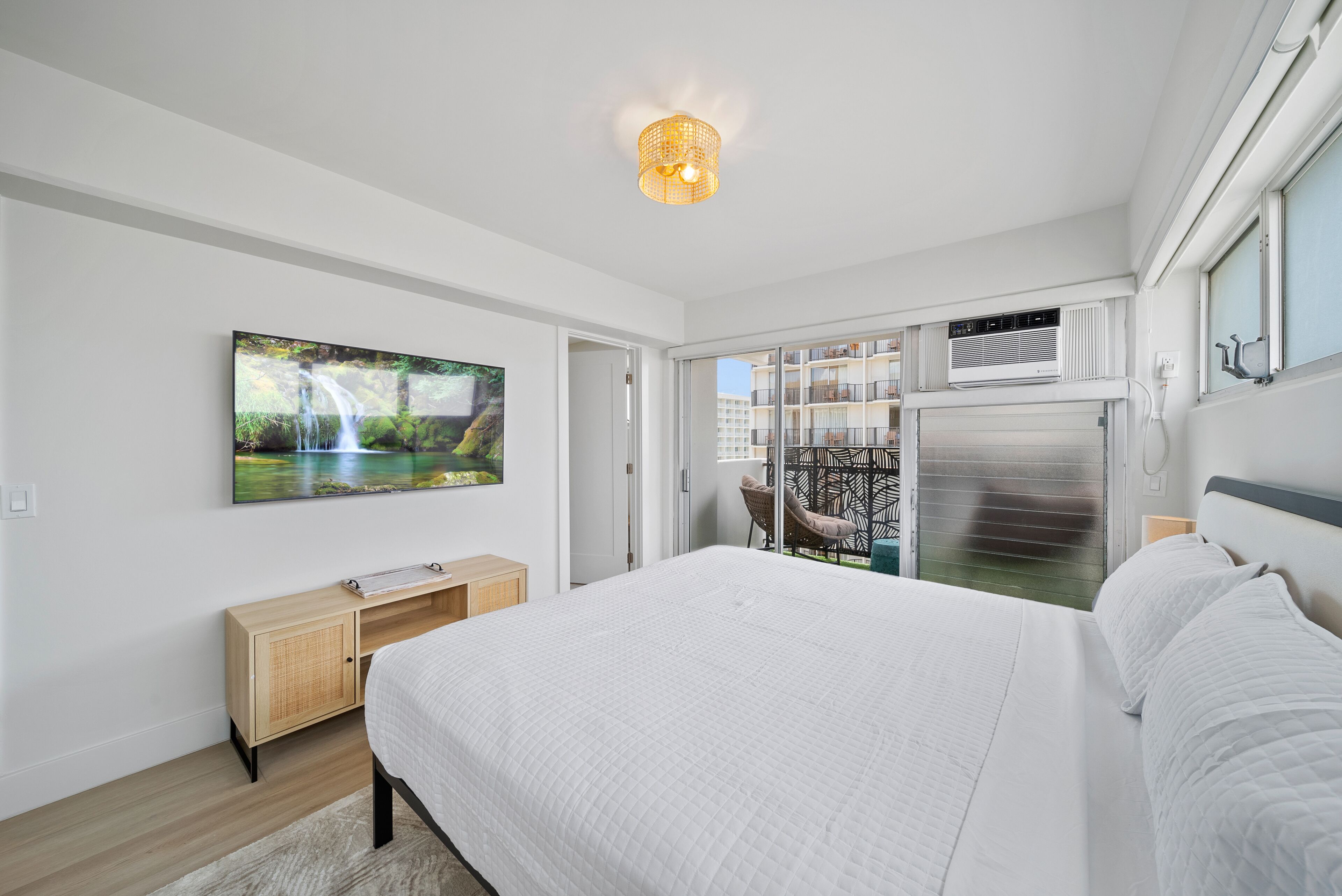 Une chambre moderne avec un grand lit, un téléviseur à écran plat, une armoire en bois et une vue sur un balcon doté d’une balustrade.