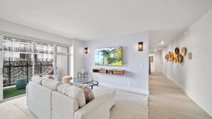 Ein modernes Wohnzimmer mit einer Couch, einem an der Wand befestigten Fernseher und Blick auf einen Balkon mit Gartenmöbeln.