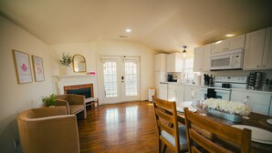 Ein geräumiges Wohnzimmer mit Kamin, eine Essecke mit Tisch für zwei Personen und eine Küche mit weißen Schränken und modernen Geräten.