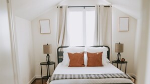 Ein Schlafzimmer mit einem Bett, Deckenventilator, zwei Nachttischen mit Lampen, einem Fenster mit Vorhängen und Holzboden.
