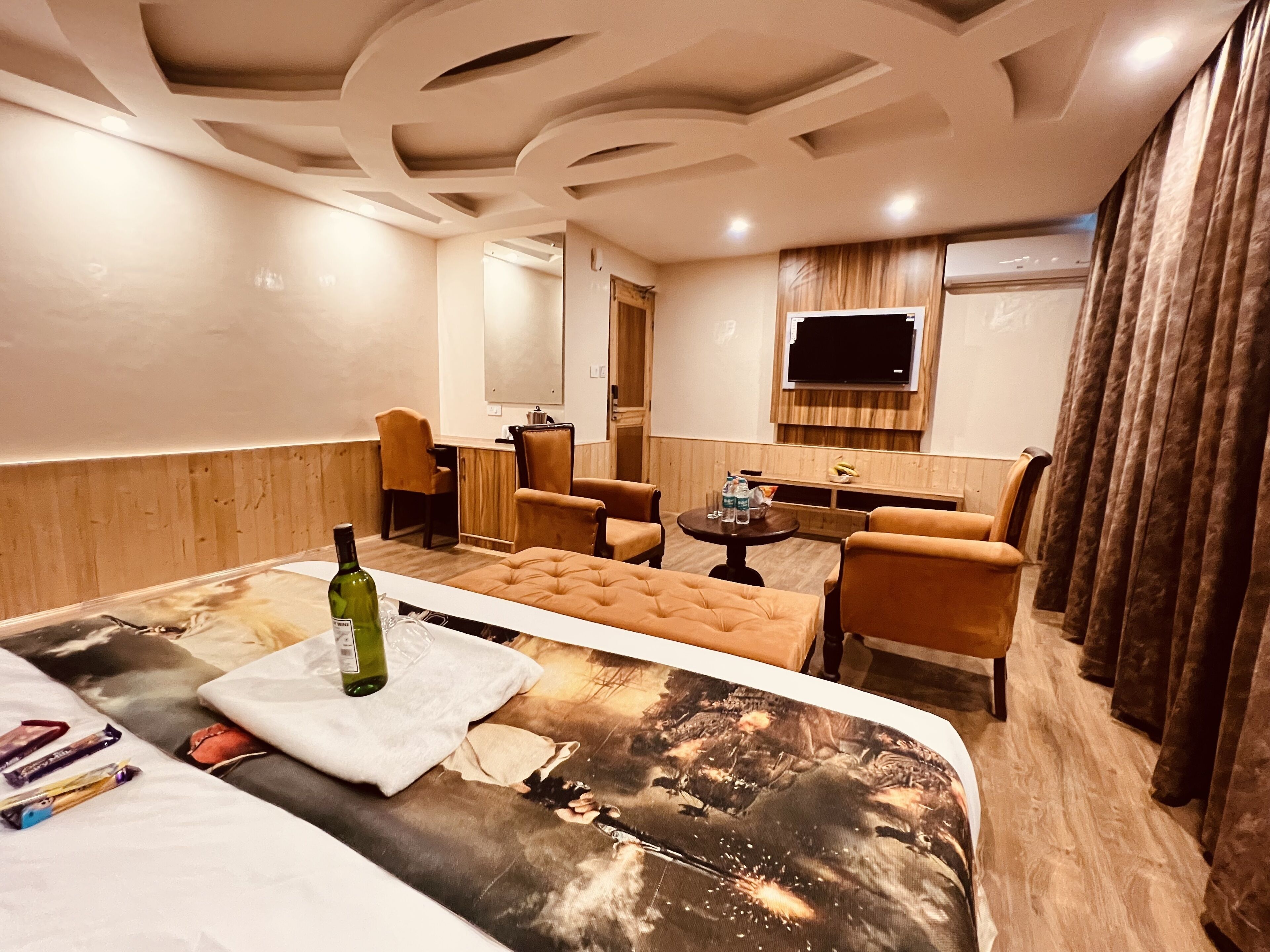 A hotel room with a large bed, a small table, two armchairs, a TV, and a wooden panel wall.