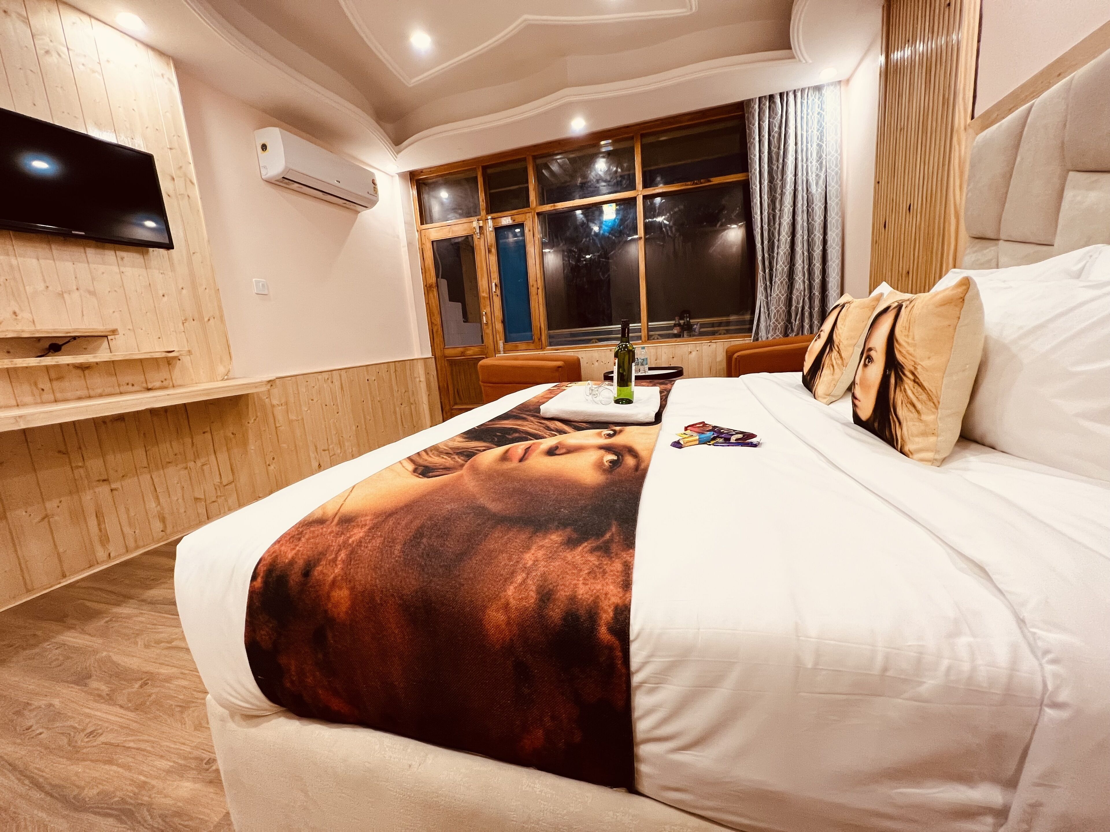 A hotel room with a bed, a TV mounted on the wall, a window with curtains, and a wooden panel wall.