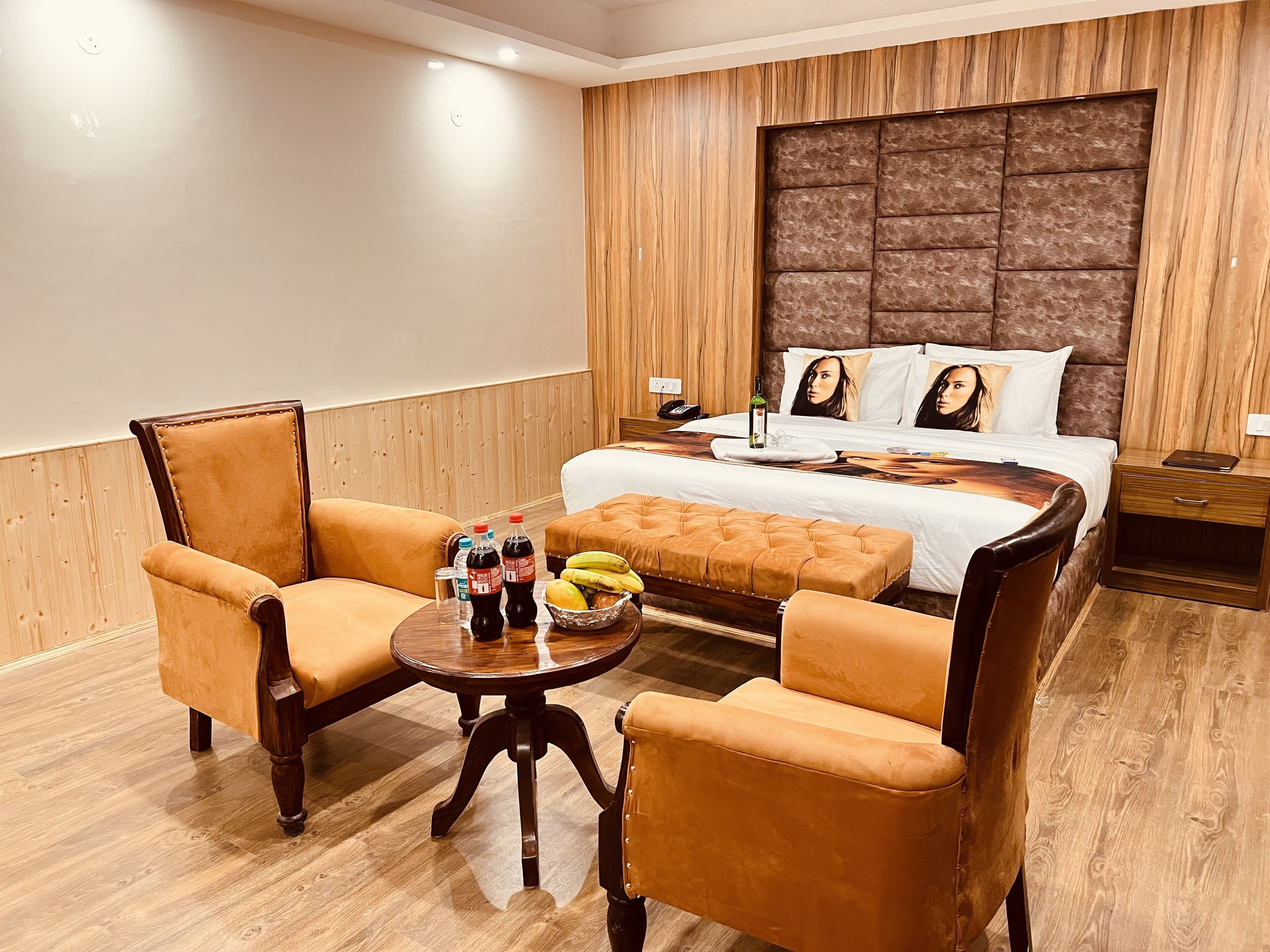 Una habitación de hotel con una cama grande, dos sillones, una mesa redonda con una botella de agua, una botella de refresco y un bol de fruta.