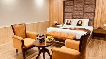Una habitación de hotel con una cama grande, dos sillones, una mesa redonda con una botella de agua, una botella de refresco y un bol de fruta.