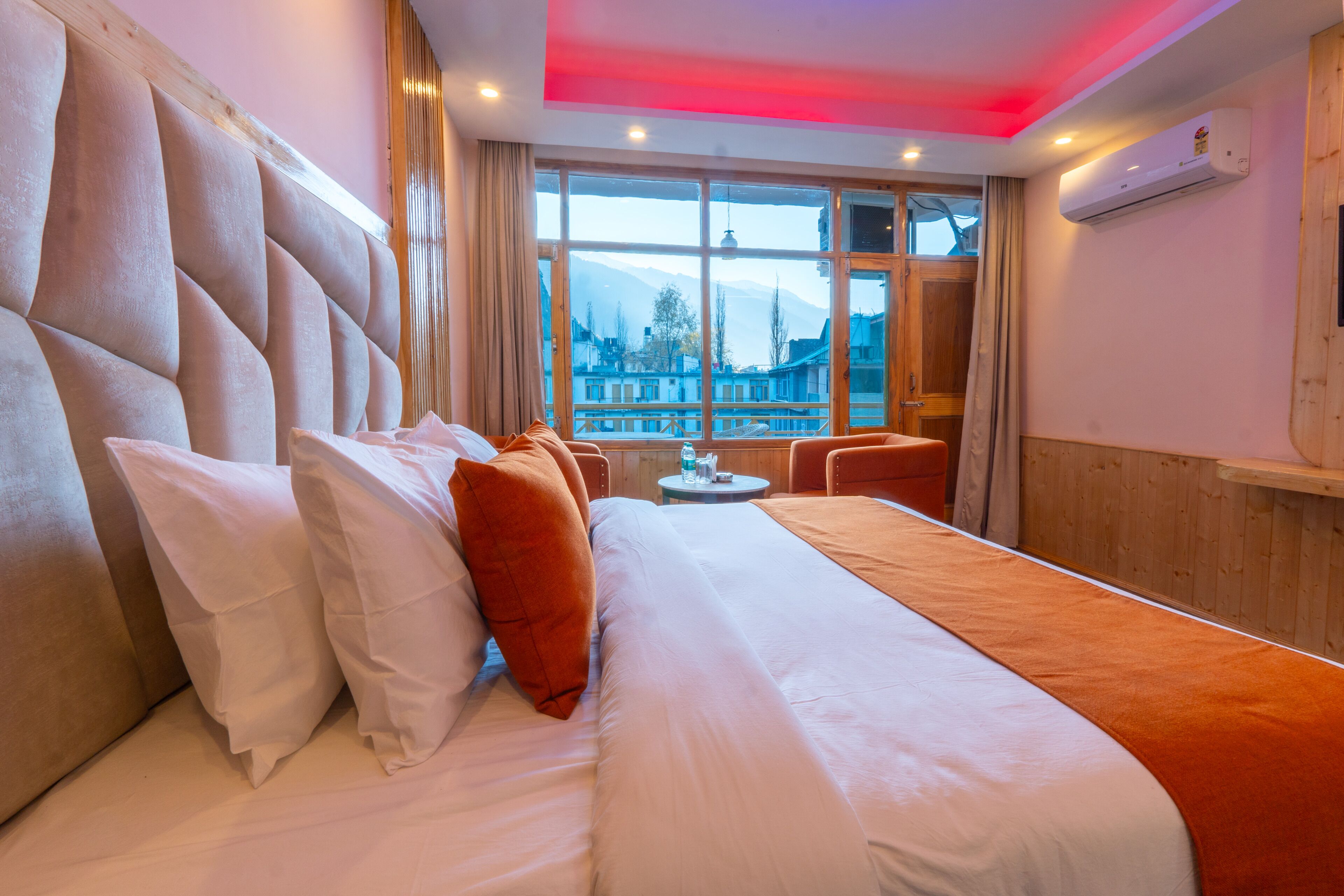 A hotel room with a large bed, a view of a cityscape through a large window, and a wooden headboard.