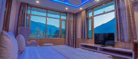 Quarto de hotel moderno com uma cama grande, televisão e vista para montanhas através de amplas janelas.