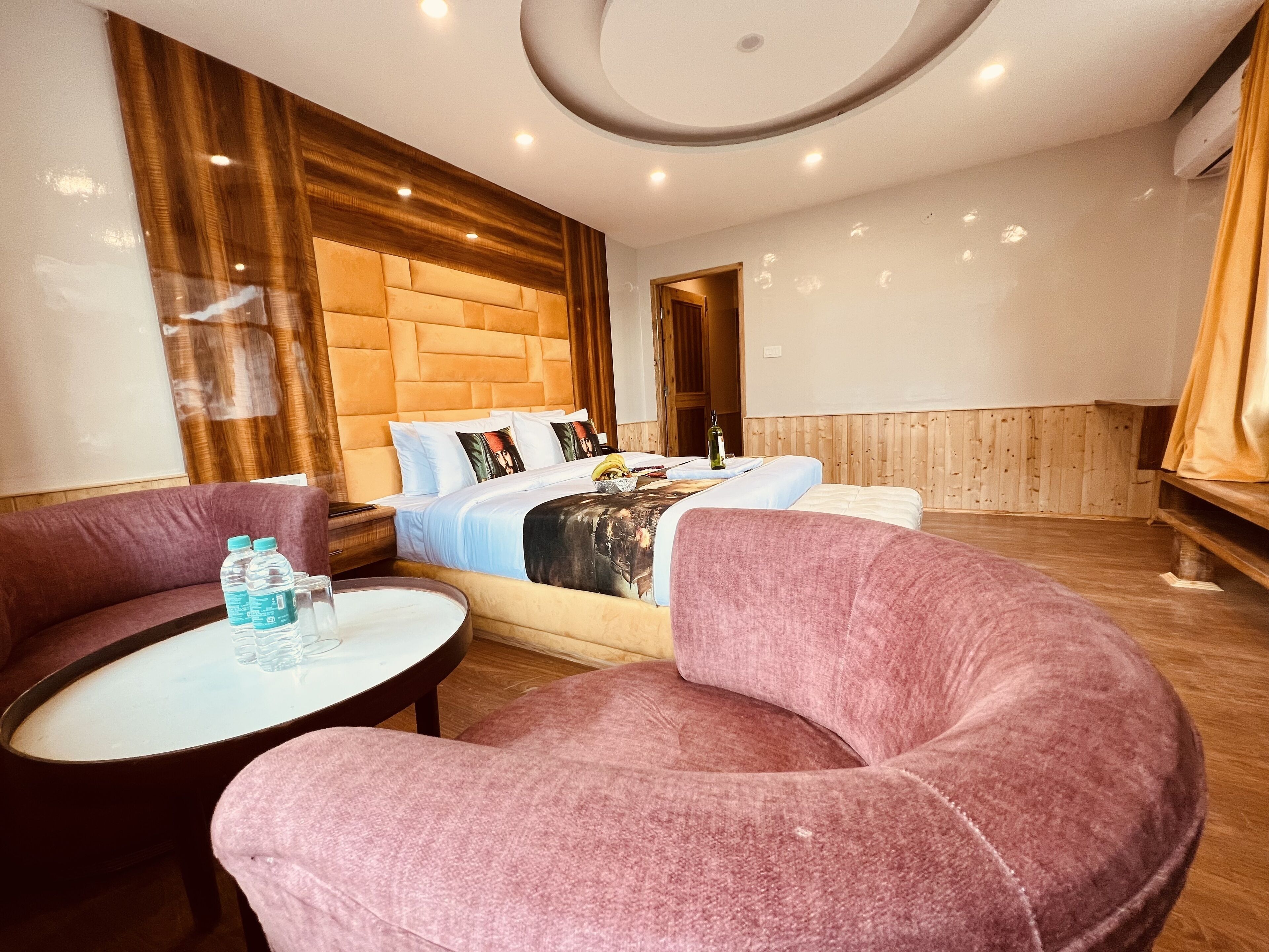 A hotel room with a large bed, two pink armchairs, a small table with water bottles, and a wooden headboard.