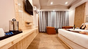 Una habitación de hotel con piso de madera, una cama, un sofá, un televisor montado en la pared, una mesita con una botella y vasos, y una ventana con cortinas.