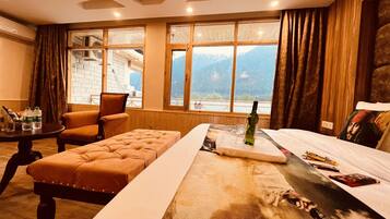A hotel room with a large bed, a wooden bench, a table with bottles, and a view of mountains through large windows.