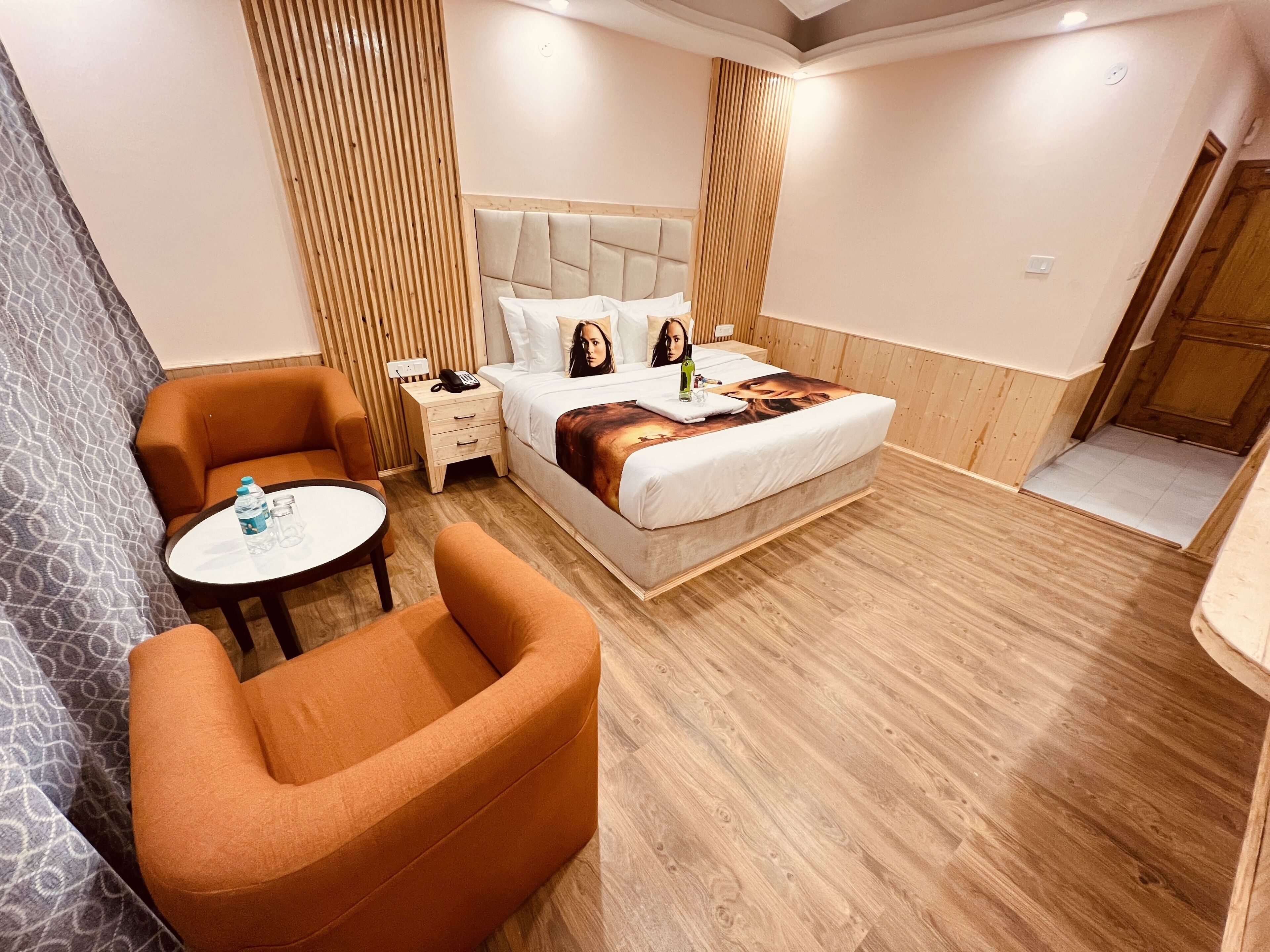 A hotel room with a bed, a round table, an orange armchair, and a wooden floor.
