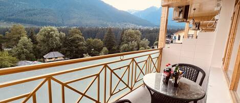 A balcony with a view of a river and mountains, featuring wicker chairs and a table with bottles.