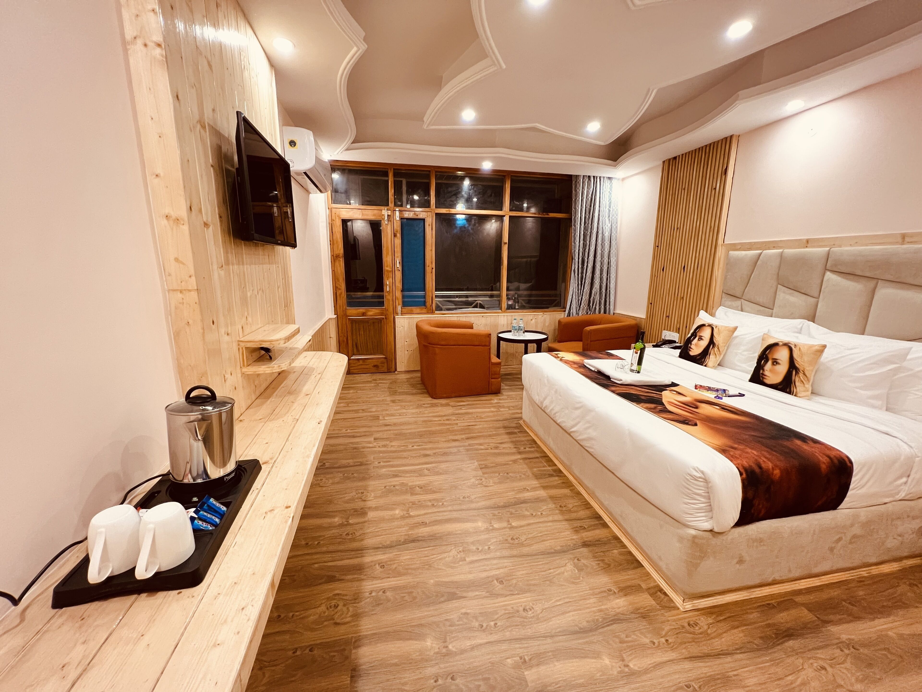 A modern hotel room with a wooden floor, a bed with a headboard, a seating area with a table, and a TV mounted on the wall.