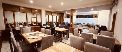A restaurant with multiple dining tables and chairs, a kitchen area, and a view of the cityscape.