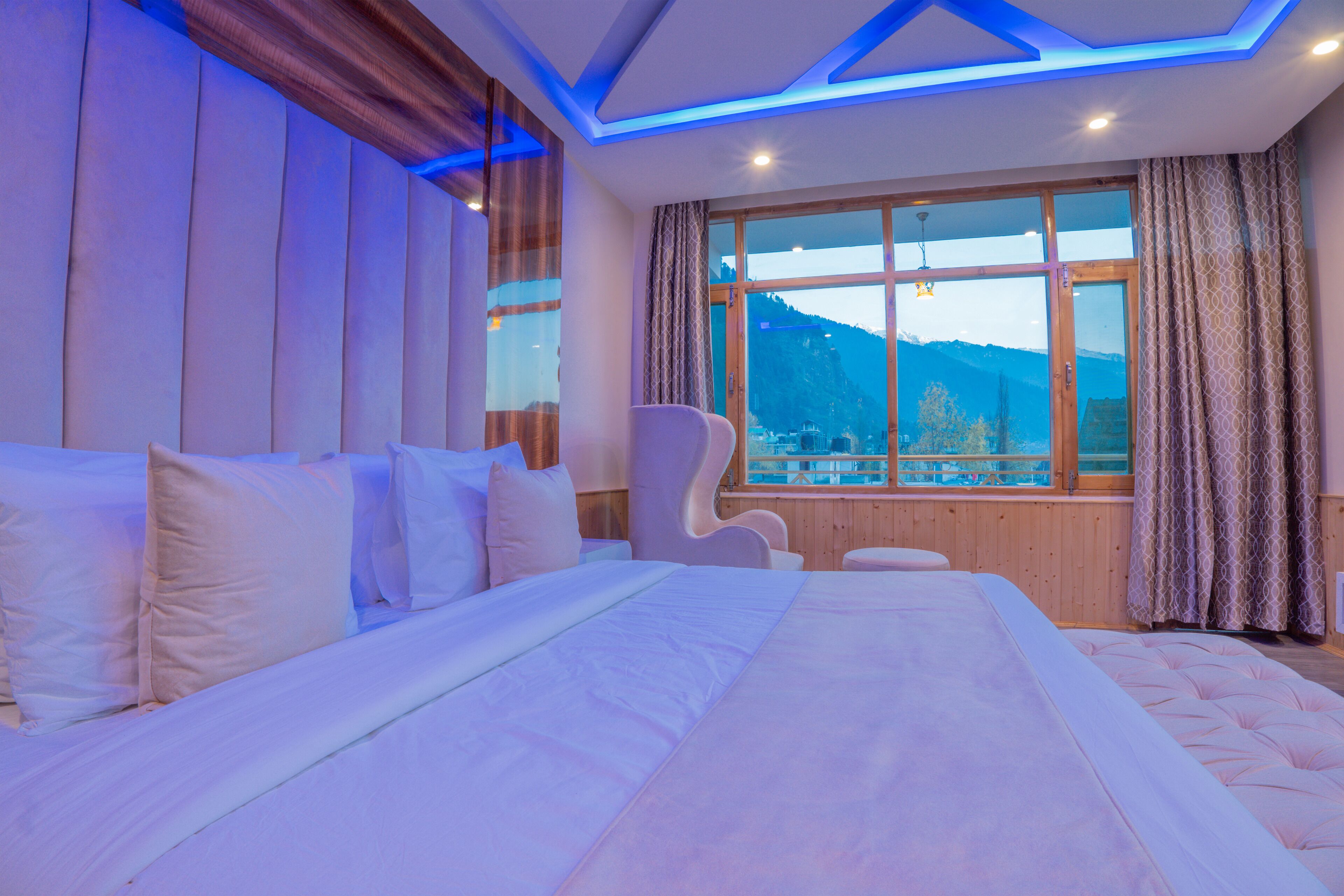 A large bed with white linens, a wooden headboard, and a view of mountains through a large window with curtains.