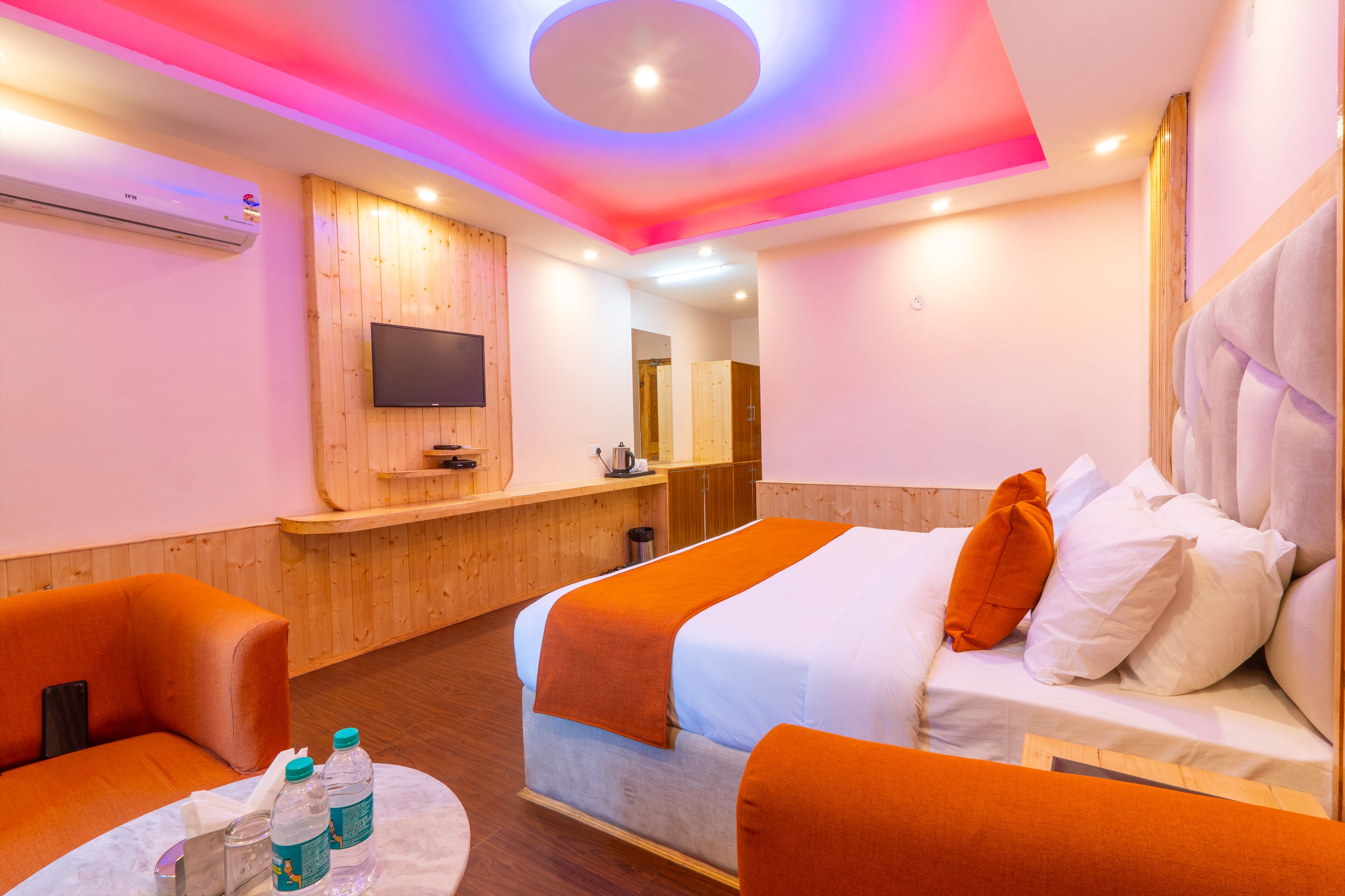 A hotel room with a bed, two orange armchairs, a small table with bottles, a TV mounted on the wall, and a wooden reception desk.